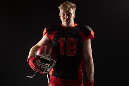 American football player standing with helmet against black background. Strong American Football Player concept for Championship Football Tournamentの写真素材