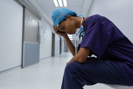 Side view of tensed young African-american female surgeon with hand on forehead sitting in the corridor at hospital. Shot in real medical hospital with doctors nurses and surgeons in authentic settingの写真素材