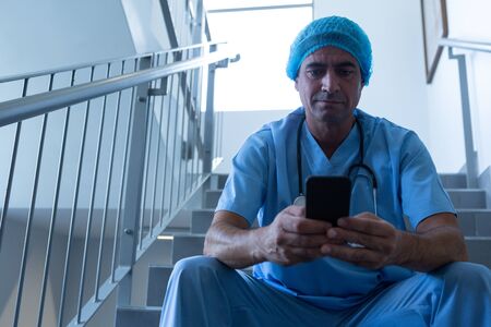 Front view of Caucasian male surgeon using mobile phone while sitting on stairs at hospital. Shot in real medical hospital with doctors nurses and surgeons in authentic settingの写真素材