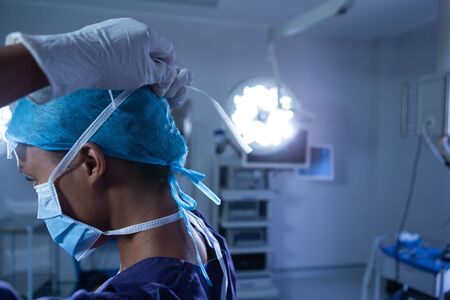 Side view of young African-american female surgeon putting surgical mask in operating room at hospital. Shot in real medical hospital with doctors nurses and surgeons in authentic settingの写真素材