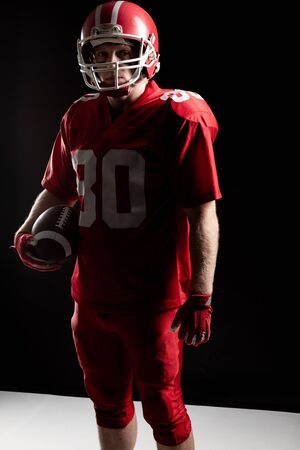American football player standing with rugby ball and helmat against black background. Strong American Football Player concept for Championship Football Tournamentの写真素材