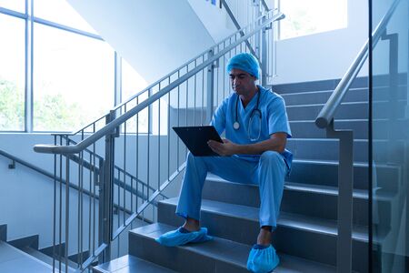 Front view of Caucasian male surgeon writing on clipboard while sitting on stairs at hospital. Shot in real medical hospital with doctors nurses and surgeons in authentic settingの写真素材