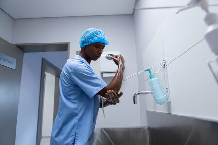 Side view of African-american male surgeon washing hands in sink  following the protocol at hospital. Shot in real medical hospital with doctors nurses and surgeons in authentic settingの写真素材
