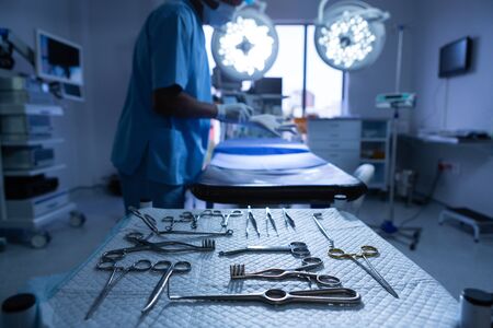 Close-up of surgical instruments arranged on table in operating room while African-american male surgeon putting gloves on background. Shot in real medical hospital with doctors nurses and surgeons in authentic settingの写真素材