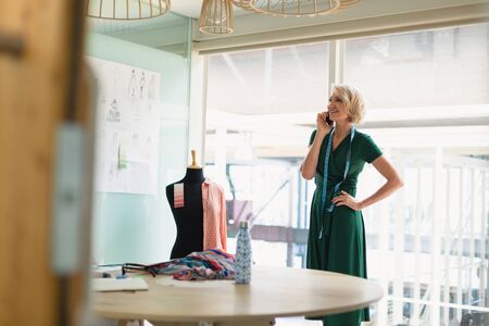 Front view of happy mature Caucasian female fashion designer talking on mobile phone in design studio. Modern casual creative business conceptの写真素材