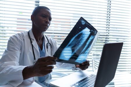 Side view of African American male doctor holding and examining x-ray report at desk in the...