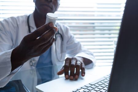 Mid section of African American male doctor holding bottle of pills in hand while using laptop in...