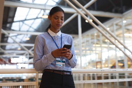 Low angle view of beautiful young mixed-race businesswoman leaning on railing and using mobile phone...