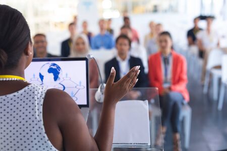 Rear view of pretty African-american female speaker giving speech to diverse people in a business seminar. International diverse corporate business partnership conceptの写真素材