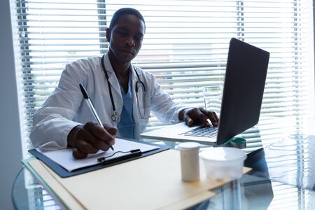 Front view of African-american male doctor writing on a clipboard with a pen while using laptop at desk in the hospital. Shot in real medical hospital with doctors nurses and surgeons in authentic settingの写真素材