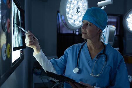 Front view of focused Caucasian female surgeon examining x-ray while writing on clipboard in operating room at hospital. Shot in real medical hospital with doctors nurses and surgeons in authentic settingの写真素材