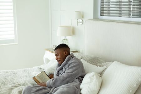 Side view of African-american man reading a book while lying on bed in bedroom at comfortable home. Authentic home lifestyle setting with young African American maleの写真素材