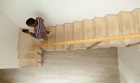 High angle aerial view of African-american man using mobile while walking down stairs in a comfortable home. Authentic home lifestyle setting with young African American maleの写真素材
