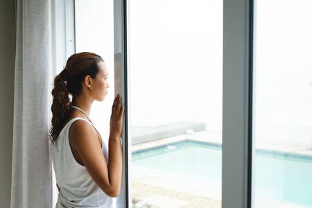 Side view of thoughtful mixed-race woman looking through window in living room at home. Summer fun...