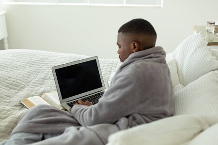 Rear view of African-american man using laptop while lying on bed in bedroom at comfortable home. Authentic home lifestyle setting with young African American maleの写真素材