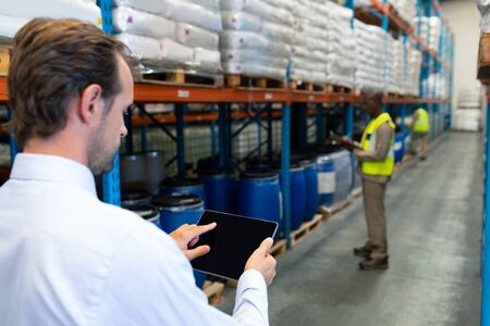 Rear view of mature Caucasian male supervisor working on digital tablet in warehouse. African-american colleagues working in front of him. This is a freight transportation and distribution warehouse. Industrial and industrial workers conceptの写真素材
