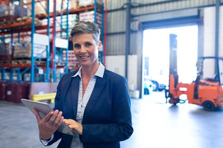 Portrait of beautiful mature Caucasian female manager looking at camera while using digital tablet in warehouse. This is a freight transportation and distribution warehouse. Industrial and industrial workers conceptの写真素材