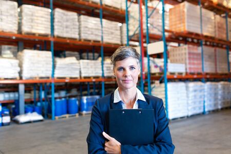 Portrait of beautiful mature Caucasian female manager holding clipboard and looking at camera in warehouse. This is a freight transportation and distribution warehouse. Industrial and industrial workers conceptの写真素材