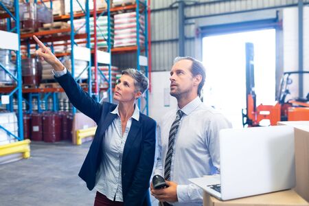 Front view of beautiful mature Caucasian female manager standing with Caucasian male supervisor and pointing at distance in warehouse. This is a freight transportation and distribution warehouse. Industrial and industrial workers conceptの写真素材