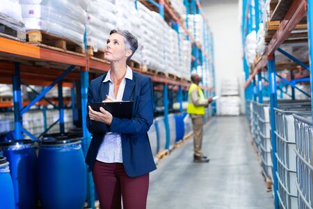 Front view of beautiful mature Caucasian female manager checking stocks on clipboard in warehouse....