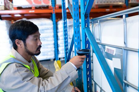 Side view of mature Asian male worker scanning package with barcode scanner in modern warehouse....