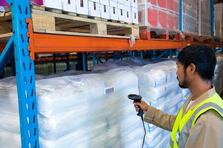 Side view of mature Asian male worker scanning package with barcode scanner in modern warehouse....