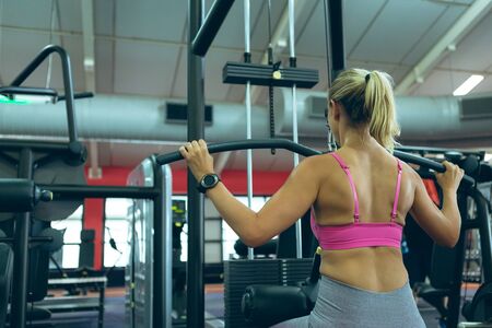 Rear view of beautiful young Caucasian female athlete exercising with lat pulldown machine in...