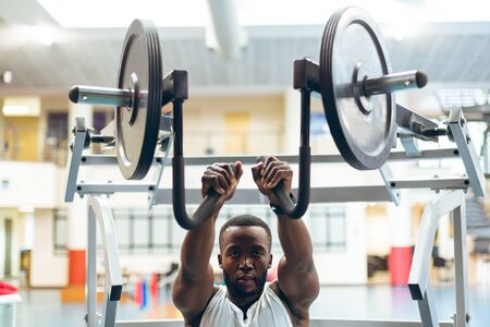 Front view of young African-american man exercising with shoulder machine in fitness studio. Bright modern gym with fit healthy people working out and trainingの写真素材