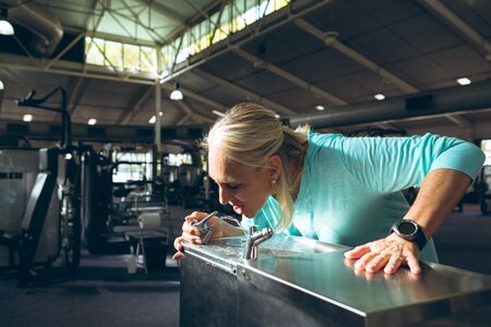 Side view of tired active senior Caucasian woman drinking tap water in fitness center. Strong active...