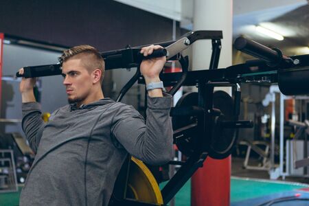 Side view of young Caucasian man exercising with shoulder machine in fitness studio. Bright modern gym with fit healthy people working out and trainingの写真素材