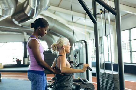Side view of young fit African-american female trainer assisting active senior Caucasian  woman on...