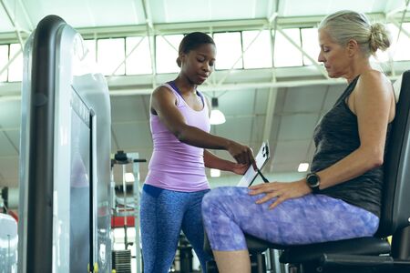 Side view of young African-american female trainer assisting active senior Caucasian woman to work...