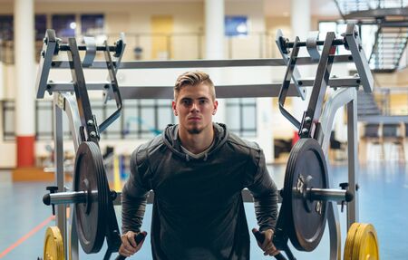 Front view of young fit Caucasian man exercising with shoulder machine in fitness studio. Bright...