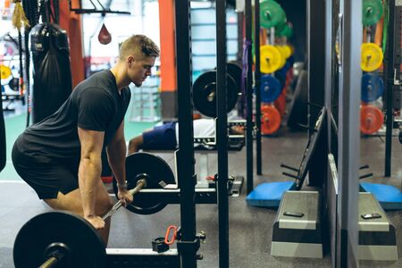 Side view of handsome fit young Caucasian male athletic exercising with barbell in fitness center. Bright modern gym with fit healthy people working out and trainingの写真素材