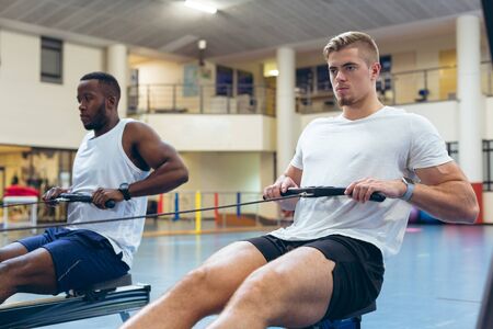 Front view of young fit diverse male athletes exercising with rowing machine in fitness studio....