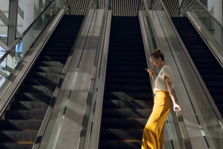 Side view of young Caucasian businesswoman using smartphone leaning on the handrail as she descends...