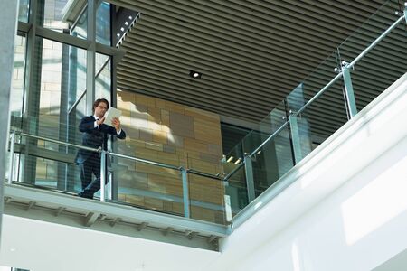 Low angle view of businessman leaning on railing and using digital tablet in corridor at modern...