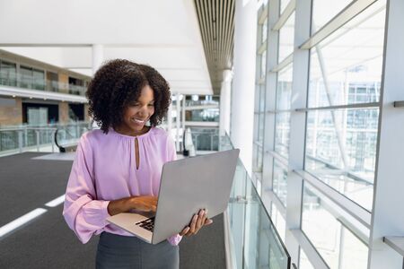 Front view of young African American businesswoman standing using laptop computer in the glass...