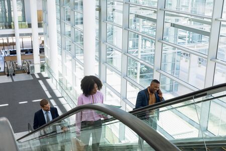 Front view of a young African American businesswoman going up an escalator in a modern building, Beside her a young African American businessman using a smartphone is going up on the next escalator. In the background a young Caucasian businessman is also coming up the escalor behind them. Modern corporate start up new business concept with entrepreneur working hardの写真素材