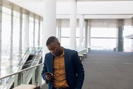 Front view of young African American businessman using smartphone in the glass walled lobby of a...