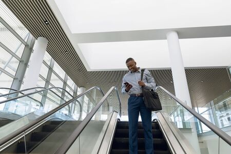 Low angle front view of smiling young African American businessman going down an escalator in a modern building using his smartphone. Modern corporate start up new business concept with entrepreneur working hardの写真素材