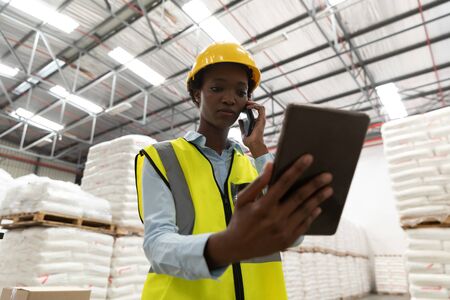 Low angle view of female worker using digital tablet while talking on mobile phone in warehouse....
