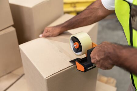 Close-up of male worker packing cardboard box with tape gun dispenser in warehouse. This is a...