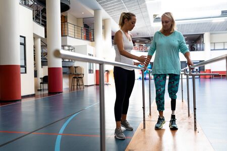 Front view of female physiotherapist helping disabled senior woman walk with parallel bars in sports...