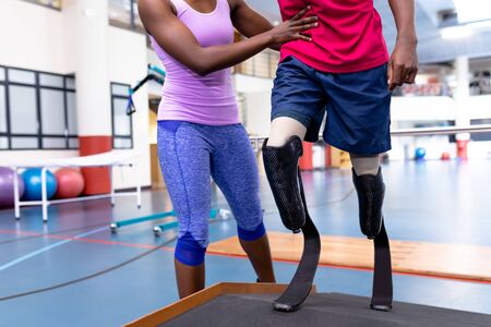 Low section of African-american female physiotherapist helping disabled African-american man walk...