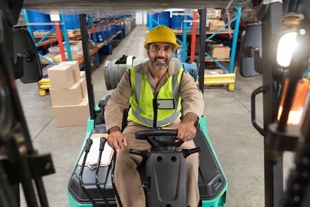 Happy male staff looking at camera while driving forklift in warehouse. This is a freight...