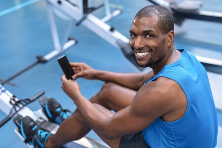 Portrait of African-american fit man using mobile phone while exercising on rowing machine in...