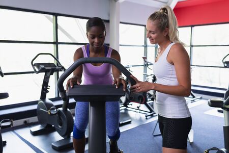 Front view of African-american Female trainer assisting woman to work out on exercise bike in...