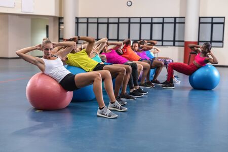 Side view of diverse female trainer training people to perform yoga on exercise ball in fitness...