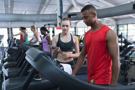 Front view of Caucasian female trainer assisting man to work out on treadmill. Bright modern gym...